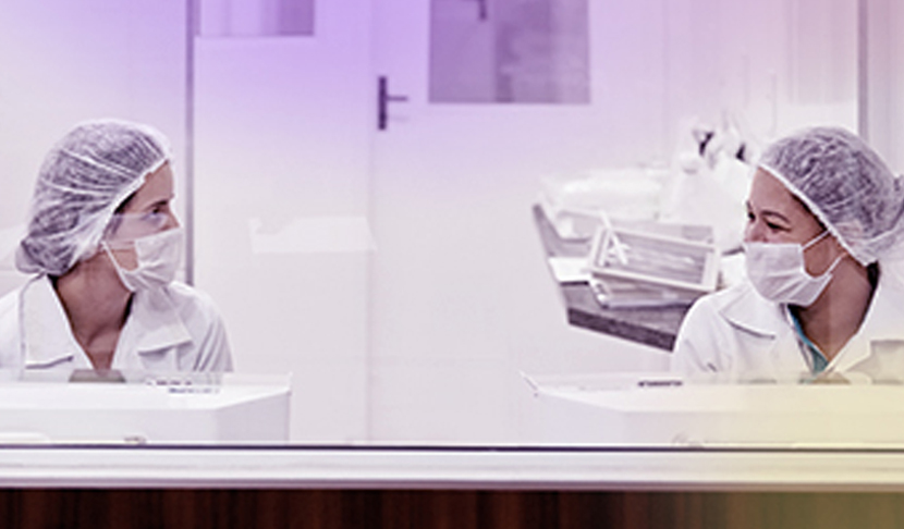 Dve laboratorijski delavki v halji, z masko in čepico na glavi, sedita vsaka za svojo napravo in se pogovarjata.